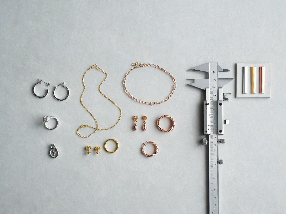 A spread of finished jewelry prototypes in different metal tones laid on a neutral work surface next to calipers and a metal sample card