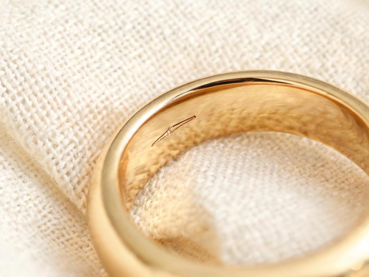 Close-up of the inner band of a gold ring showing a subtle laser-engraved private label mark, resting on a neutral linen surface with soft natural light.